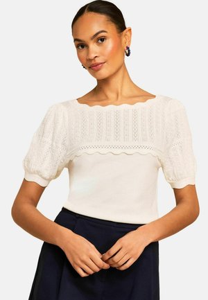 Femeie purtând un top alb tricotat cu mâneci bufante și margini zimțate, asortat cu pantaloni închiși la culoare cu talie înaltă, stând cu mâinile împreunate.