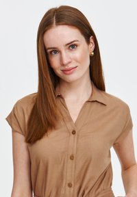 Beżowa koszula zapinana na guziki z krótkimi rękawami, z kołnierzykiem i luźnym krojem. Teksturowana tkanina z drewnianymi guzikami dodaje szczegółów.