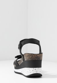 Sandale compensée noire avec bride cheville réglable, semelle intérieure en liège texturé, et semelle tachetée blanche et noire vue de dos.