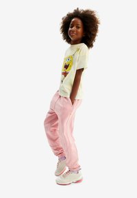 Enfant portant un t-shirt tie-dye clair avec un motif SpongeBob, un pantalon de survêtement rose avec des coutures latérales, et des chaussures blanches avec des accents roses.