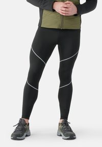 Sorte sportsleggings med refleksive sølvaccentlinjer, parret med grønne og grå teksturerede sneakers, designet til ydeevne og komfort.