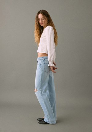 Blusa bianca corta con maniche lunghe, jeans strappati a vita alta blu e scarpe nere slip-on con borchie; modello in posizione laterale.