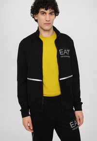 Black zip-up jacket with reflective accent, worn over a bright yellow shirt. The jacket features a logo on the left chest area and elastic cuffs.