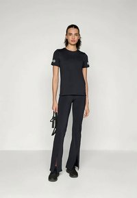 Sieviete stāv pret vienkāršu fonu, uzvilkusi melnas bikses ar šķeltu apakšmalu, melnu īsām piedurknēm augšu, melnas kurpes un tur rokās mazu melnu somiņu.