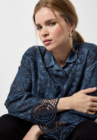 Camisa texturizada azul oscuro con detalles florales. Las mangas largas presentan un intrincado borde de encaje. Diseño con cuello y botones. Fondo negro.
