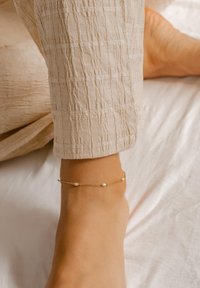 Złota bransoletka na kostkę z małymi białymi koralikami, charakteryzująca się delikatnym wzorem łańcuszka. Zbliżenie na stopę na tle teksturowanej beżowej tkaniny.