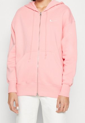 Osoba nosząca jasnoróżową bluzę Nike na zamek z sznurkami i białe spodnie, stojąca na jednolitym tle.