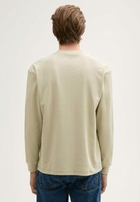 Długi rękaw, jasnobeżowa bluzka o teksturowanej tkaninie. Posiada okrągły dekolt i ściągacze przy rękawach. Stylizowana z niebieskimi dżinsami.