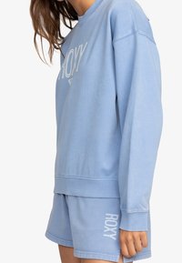 Felpa azzurra chiaro e pantaloni corti abbinati con la scritta bianca "ROXY". La felpa presenta polsini e orlo a coste, con una texture in cotone morbido.