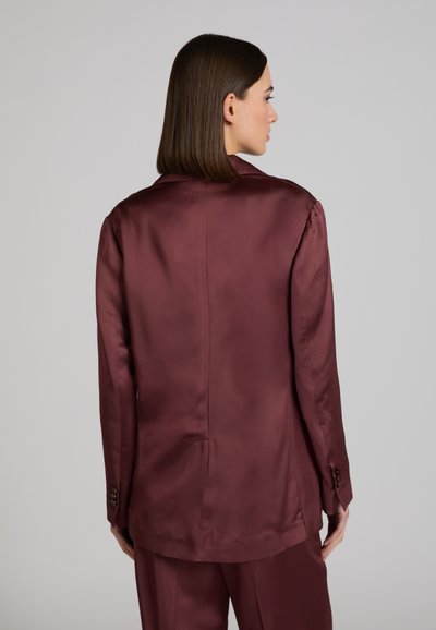Frau mit glattem braunem Haar, die abgewandt ist und einen lockeren, glänzenden burgunderfarbenen Blazer sowie passende Hosen vor einem schlichten grauen Hintergrund trägt.