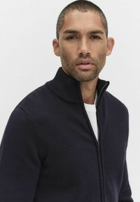 Granatowy sweter zapinany na zamek z prążkowanym kołnierzem, wykonany z teksturowanej dzianiny i dopasowany do sylwetki, noszony na białej koszulce z okrągłym dekoltem.