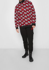 Sudadera con capucha negra con un patrón audaz y repetitivo del logo "HUGO" en rojo y blanco, combinada con pantalones de chándal negros con bolsillo lateral tipo cargo.