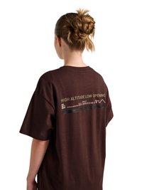 Femme avec un chignon tressé portant un t-shirt ample marron foncé avec le texte "HIGH ALTITUDE LOW OPENING" dans le dos.
