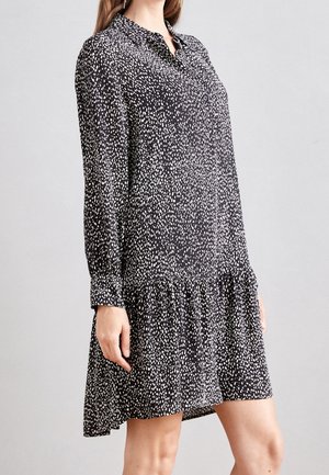 Kobieta ubrana w długą czarną sukienkę z nieregularnym białym wzorem w kropki, z przodu zapinaną na guziki, z falbaną na dole, stojąca na jednolitym tle.