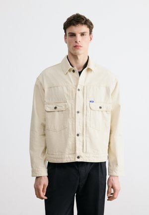 Bărbat tânăr purtând o jachetă crem cu nasturi și buzunare frontale, și pantaloni negri, stând într-un fundal alb simplu.