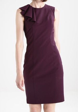 Robe sans manches de couleur violet foncé, arrivant au genou, avec un détail en volants à l'épaule gauche, portée par une personne debout sur un fond blanc uni.