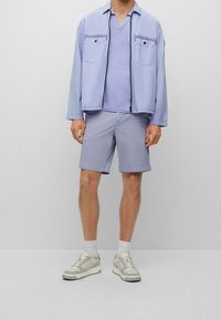 Man wearing light purple shorts, a matching T-shirt, a light jacket, white socks, and white sneakers standing against a plain wall.