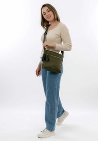 Green leather crossbody bag with a tassel accent, silver zipper, and adjustable strap. The model wears casual blue jeans and white sneakers.