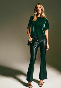 Haut en velours vert avec bord en dentelle, associé à un pantalon évasé en simili cuir noir et des sandales noires à lanières. Un petit sac bandoulière complète l'ensemble.