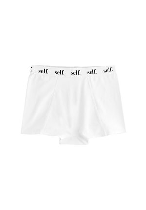 Vrouw draagt witte sportshorts en sportbeha met "self." gedrukt op de elastische banden, poseert met hand bij de tailleband.