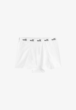 Vrouw draagt witte sportshorts en sportbeha met "self." gedrukt op de elastische banden, poseert met hand bij de tailleband.