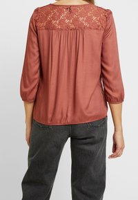 Blusa color ruggine con pannello in pizzo sul retro, maniche a tre quarti e una vestibilità leggermente ampia con dettagli arricciati.