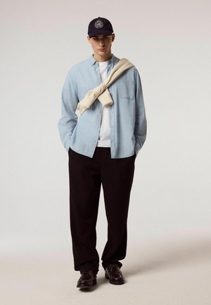 Młody mężczyzna ubrany w jasnoniebieską koszulę na guziki nałożoną na białą koszulkę, beżowy sweter zawiązany na ramionach, czarne spodnie, czapkę i brązowe buty, stojący.