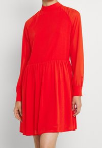 Punainen mekko korkealla kaula-aukolla, pitkät raglan-hihat ja kootun vyötärön. Valmistettu kevyestä kankaasta, jossa on virtaava hame.