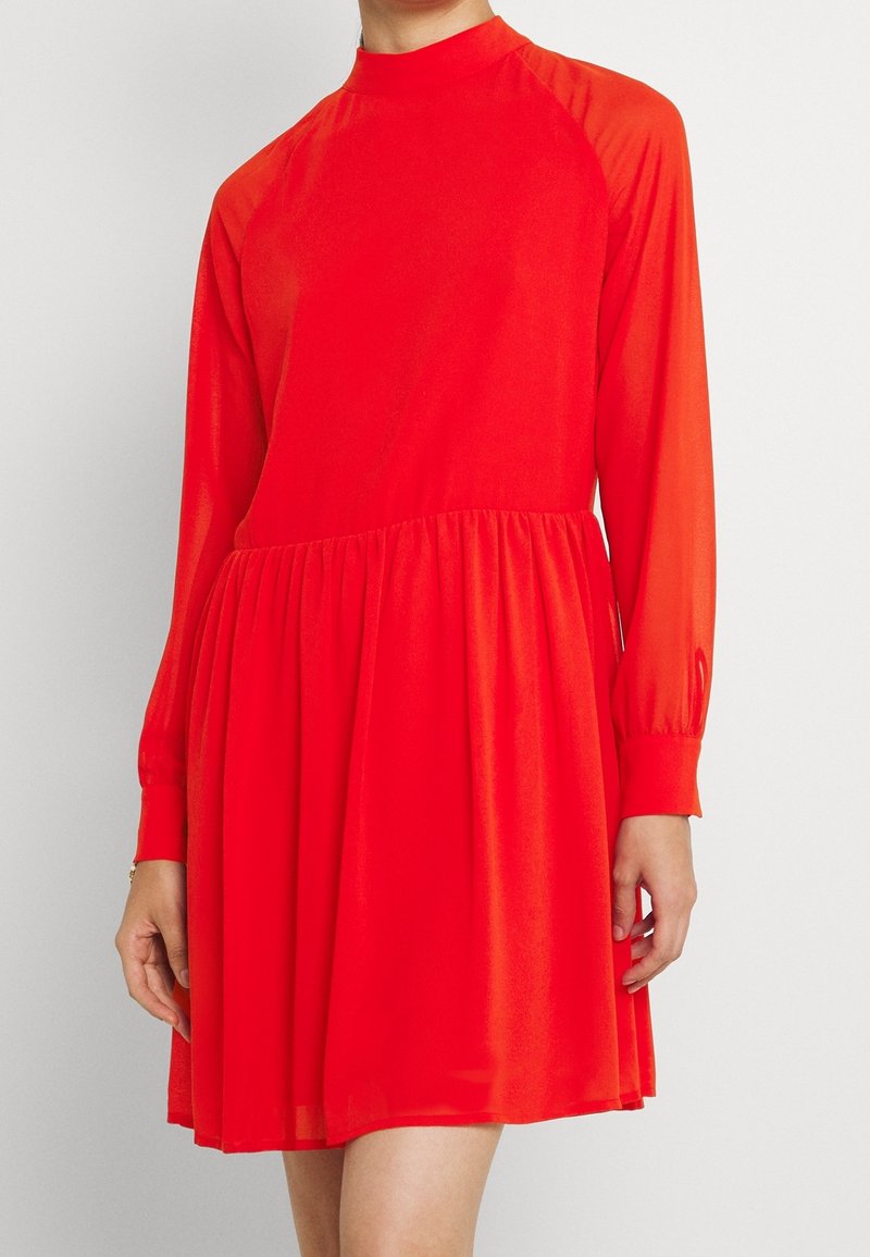 Punainen mekko korkealla kaula-aukolla, pitkät raglan-hihat ja kootun vyötärön. Valmistettu kevyestä kankaasta, jossa on virtaava hame.