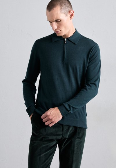 Moški v temno zeleni dolgi rokavi z zip-up polo majico in ujemajočimi se žametnimi hlačami, gleda navzdol z eno roko v žepu in drugo, ki drži rob majice.