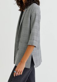 Blazer à carreaux gris à manches courtes, avec une fermeture à un bouton et des fentes latérales. Fabriqué dans un tissu lisse avec un motif texturé.