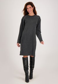 Vrouw in een langwerpige, donkergrijze jurk tot de knie met studdetails op de schouders en zwarte kniehoge laarzen, lachend en staand.