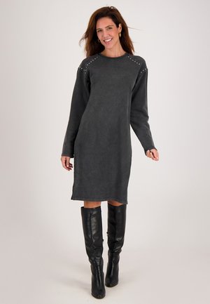 Vrouw in een langwerpige, donkergrijze jurk tot de knie met studdetails op de schouders en zwarte kniehoge laarzen, lachend en staand.
