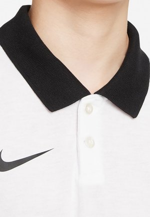 Polo shirt blanc avec un col noir, comportant trois boutons en nacre et un logo Nike en swoosh noir sur le côté gauche. Texture de tissu douce.