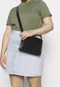Borsa a tracolla nera con motivo a coccodrillo testurizzato, forma rettangolare, chiusura con zip e tracolla regolabile, tenuta contro una gonna in denim chiaro.