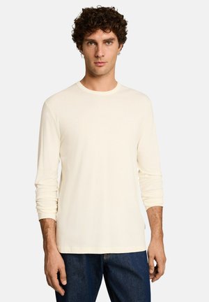 Man met krullend donker haar, gekleed in een crèmekleurig shirt met lange mouwen en donkerblauwe spijkerbroek, staand tegen een effen lichte achtergrond.