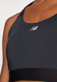 Soutien-gorge de sport noir avec une texture lisse, présentant un col rond et une large bande noire à la base, ornée d'un logo blanc.