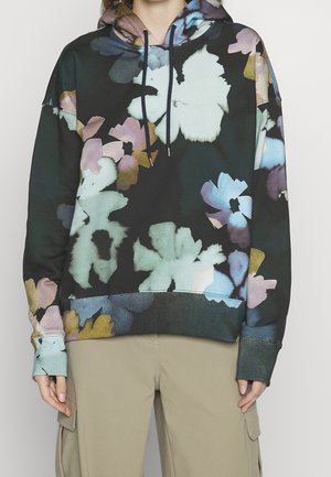 Personne portant un sweat à capuche noir avec de grandes impressions florales pastel en bleu, vert, violet et beige, associé à un pantalon cargo beige.