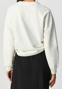 Pullover oversize crema con collo rotondo, polsini a coste e orlo posteriore corto. Abbinato a una gonna nera, presenta una morbida texture in cotone.