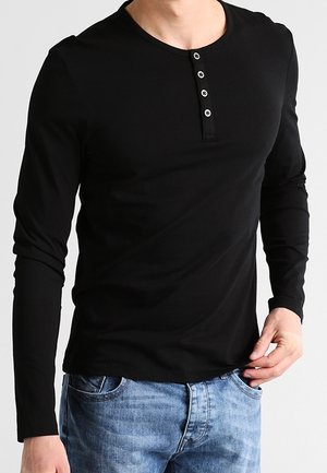Mężczyzna w czarnej koszulce henley z długim rękawem i trzema guzikami oraz jasnoniebieskich dżinsach, ręka poprawia dół koszuli po prawej stronie.