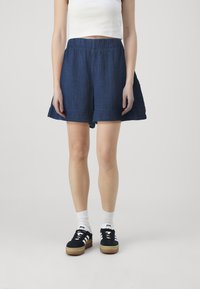 Tamsiai mėlynos, lengvos šortai su plačiu kirpimu, elastine juosta ir tekstūruotu audiniu; derinami su juodais sportbačiais ir baltais kojinėmis.