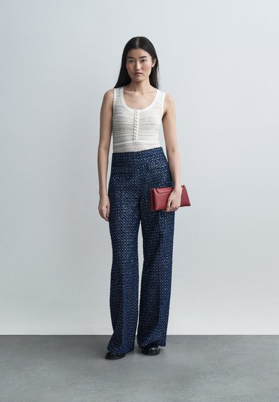 Νεαρή γυναίκα που φοράει λευκό αμάνικο πλεκτό τοπ και παντελόνι με φαρδιά μπατζάκια, σε ψηλή μέση και μπλε-σκούρο μοτίβο, κρατώντας κόκκινο clutch, στέκεται μπροστά σε μια απλή τοίχο.