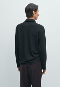 Svart stickad långärmad polo skjorta med avslappnad passform, med krage och subtil textur, bärs tillsammans med mörka byxor.