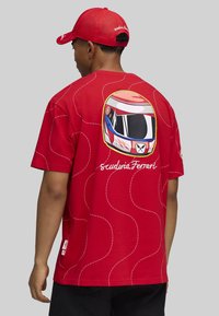Mand iført rød Scuderia Ferrari kasket og rød t-shirt med et racinghjelm grafik og "scuderia Ferrari" tekst på ryggen.