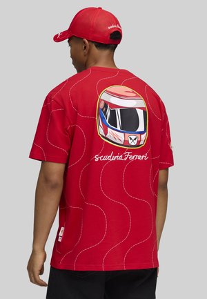 Mann trägt eine rote Scuderia Ferrari Kappe und ein rotes T-Shirt mit einem Grafikdesign eines Rennhelms und dem Text "Scuderia Ferrari" auf der Rückseite.