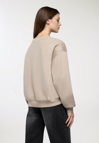 Beżowa bluza z okrągłym dekoltem, opadającymi ramionami i ściągaczami. W połączeniu z czarnymi dżinsami, które mają subtelną teksturę i minimalne detale.