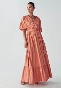 Persikka värinen, kerroksittain leikatun maksi mekon, jossa on kietaisumalli, lyhyet puffihihat ja yhteensopiva vyö vyötäröllä. Silkkinen kankaan pinta.