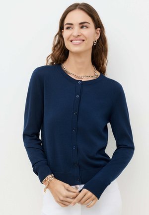 Nainen pukeutuneena tummansiniseen napitettuun neuletakkiin, valkoisiin housuihin sekä kultaisen ketjun kaulakoruun, rannekoruun ja korvakoruihin, hymyilee ja katsoo sivulle.