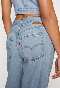 Světle modré džíny s vysokým pasem, s koženým štítkem na zadní straně, dvěma zadními kapsami a oranžovým prošíváním.