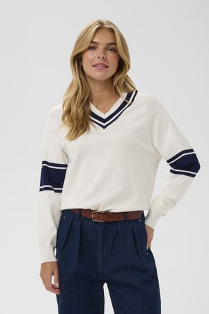Pull blanc avec des accents de rayures bleu marine sur les manches et le col, design en V, associé à un pantalon en denim foncé taille haute et une ceinture marron.